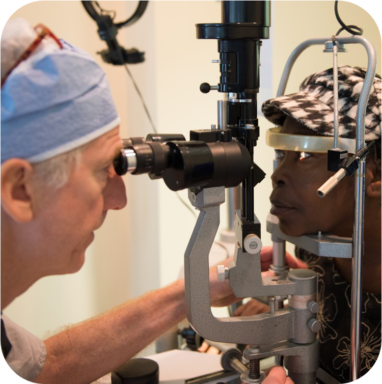 一名眼睛上绑着纱布的小孩正趴在一名妇女的肩头休息，妇女穿着格子衬衫，抱着他穿过一家医疗机构的狭窄走廊，走廊上还能看到一名穿手术服的医疗工作者。这场景表明小孩刚刚接受了眼部手术，正处于术后护理或恢复阶段。